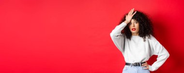 Sinirli ve yorgun genç bir kadın alnını tokatlıyor, facepalm yapıyor ve kameraya bakıyor şaşkın şaşkın, kırmızı arka planda duruyor..