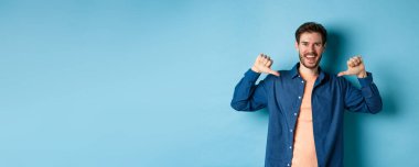 Kendi reklamını yapan mutlu genç adam, kendini işaret ediyor ve gülümsüyor, mavi arka planda profesyonel gibi duruyor..