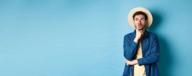 Düşünceli erkek turist yaz tatilini düşünüyor, düşünceli bir yüz ifadesiyle bir kenara bakıyor, dudaklarına dokunuyor ve bir sonraki seyahati düşünüyor, mavi arka planda duruyor..