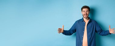 Mutlu gülümseyen adam ellerini yana açıp büyük bir nesne gösteriyor. Büyük nesnelerden bahsediyor. Mavi arka planda neşeli duruyor..