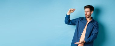 Şok olmuş genç adam boş alana bakıyor ve mavi arka planda duran büyük boyutlu nesneleri gösteriyor..