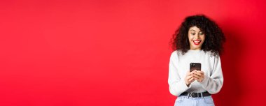 Kıvırcık saçlı, akıllı telefondan mesaj okuyan, mutlu gülümseyen, internette iyi haberler alan kırmızı arka planda duran genç bir kadın görüntüsü..