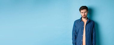 Üzgün ve kasvetli genç adam somurtuyor, sefil yüzünü çekiyor ve kameraya bakıyor, mavi arka planda duruyor..