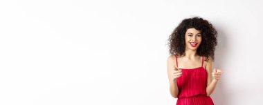 Kırmızı elbiseli güzel kadın seni tebrik ediyor, kamerayı işaret ediyor ve gülümsüyor, beyaz arka planda duruyor..