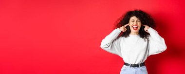 Sinirli kıvırcık kız parmaklarıyla kulaklarını tıkıyor ve bağırıyor. Kırmızı arka planda duran korkunç gürültüden rahatsız oluyor..