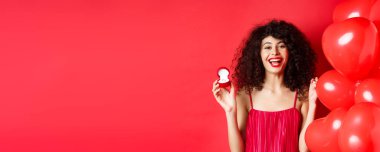 Mutlu güzel bayan evlilik teklifi için heyecanlanıyor, nişan yüzüğünü gösteriyor ve gülüyor, kırmızı arka planda kalp balonlarının yanında duruyor..