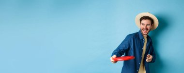 Mutlu adam gülüyor ve frizbi oynuyor, arkadaşına atıyor ve gülümsüyor, mavi arka planda yaz şapkası takıyor..