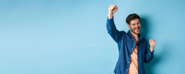 Şanslı adam zaferi kutluyor, tezahürat yapıyor ve sevinçle evet diyor, şampiyon gibi elini kaldırıyor, mavi arka planda duruyor..