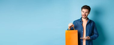 Turuncu alışveriş çantası gösteren, gülümseyen, satın alınmış, mavi arka planda duran yakışıklı beyaz adam. Boşluğu kopyala