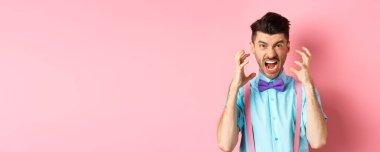 Frustrated young man losing temper, screaming and looking angry at camera, shaking hands pissed-off, standing mad on pink background.