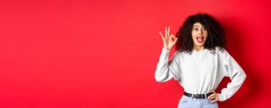 Heyecanlı güzel bir kadın evet diyor, iyi bir jest yapıyor ve hevesli görünüyor. Katılıyorum. İltifat ediyor, kırmızı arka planda duruyor..