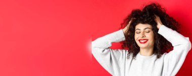 Yumuşak kıvırcık saçlara dokunan, gülümseyen, kırmızı arka planda duran kaygısız kadının yakın çekimi. Saç bakımı ve güzellik konsepti.