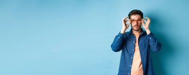 Yakışıklı genç adam mağazada yeni gözlük deniyor, kameraya bakıyor, mavi arka planda duruyor..