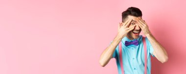 İlgi çekici papyonlu komik adam, parmaklarının arasından ilginç bir şeye bakıyor, havalı bir şeye bakıyor, pembe arka planda duruyor..