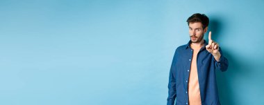 Genç bir adamın görüntüsü uyarı veriyor, ders veriyor, azarlamak için parmağını kaldırıyor, kameraya bakıyor, birine patronluk taslıyor, mavi arka planda duruyor..