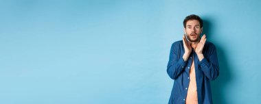 Şok olmuş ve korkmuş genç adam sersemlemiş, ellerini kaldırıyor ve nefes nefese, mavi arka planda duruyor..