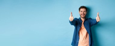 Yakışıklı, gülümseyen ve onay veren genç adam, mutlu bir yüzle evet de, mavi arka planda dur..