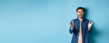 Neşeli genç adam kameraya el kaldırıyor ve gülümsüyor, senin için seviniyor, iyi iş çıkarıyor, mavi arka planda duruyor..