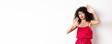 Kıvırcık saçlı ve kırmızı elbiseli genç bir kadın, durmak istiyor, birini engelliyor, savunmaya geçiyor, kendini koruyor, beyaz arka plana karşı duruyor..