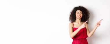 Şık kırmızı elbiseli neşeli bir kadın model, gülümsüyor ve parmaklarını logoya doğrultuyor, beyaz arka planda duruyor..