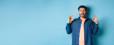 Etkileyici bir adam harika bir şeyi övüyor, iyi işaretler gösteriyor ve şaşırmış görünüyor, iltifat ediyor, mavi arka planda duruyor..