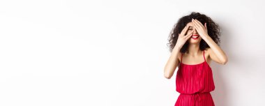 Kıvırcık saçlı, kırmızı elbiseli, elleriyle gözlerini kapatan ama parmaklarının arasından bakıp gülümseyen, ilginç, beyaz bir arka plana bakan komik bir genç kadın..