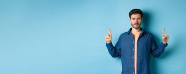 Ciddi bir beyaz adam, sakallı, boş alanı işaret eden, logoyu gösteren ve kameraya bakan, mavi arka planda duran..