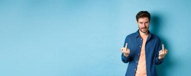 Ignorant and unbothered man showing middle-fingers and looking reluctant at camera, give zero fucks, standing on blue background.