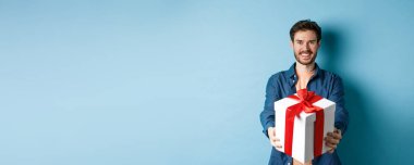 Sevgililer Günü. Yakışıklı, gülümseyen, hediye kutusuyla elini uzatan, mutlu tatiller dileyen adam. Sürpriz hediyeler hazırlayan, neşeli görünen, mavi arka planda duran bir adam..