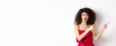 Şaşkın ve üzgün, kıvırcık saçlı, gece elbisesi ve makyajı olan, parmaklarını sağa doğru gösteren, üzgün, beyaz arka planda duran bir kadın..