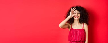 Güzel kadın heyecanla işaret ver, tanıtım teklifini kontrol et, kırmızı arka planda kırmızı renkte dur..