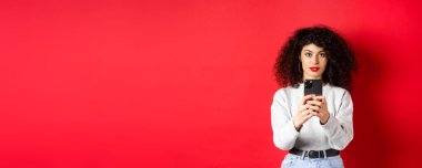 Kıvırcık saçlı, akıllı telefonuyla video kaydeden, cep telefonuyla fotoğraf çeken ve kırmızı arka planda duran genç bir kadın..