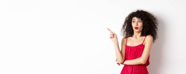 Kıvırcık saçlı heyecanlı kız, kırmızı elbise giyiyor, parmağını sola gösteriyor ve şaşırmış görünüyor, havalı reklamlar yapıyor, beyaz arka planda duruyor..