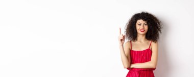 Kırmızı elbiseli güzel, gülümseyen bayan bir numarayı gösteriyor, parmağını kaldırıyor ve mutlu görünüyor, beyaz arka planda duruyor..