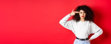 Kıvırcık saçlı, kafasında silah işareti olan, kafası patlayan, kırmızı arka planda duran, rahatsız bir beyaz kadın..