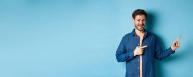 Sakallı neşeli beyaz adam boş bir yeri işaret ediyor ve kameraya gülümsüyor. Reklam yapıyor, mavi arka plan..