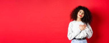 Kıvırcık saçlı, yumuşak bir genç kadın, kalbe sarılmış ve mutlu bir ifadeyle gülümsüyor, kırmızı arka planda dikilen, teşekkür edin..