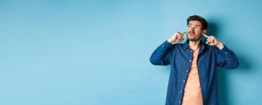 Sinirli genç adam sesi, parmaklarıyla kulaklarını kapatmış ve yüzünü buruşturmuş, gürültücü komşular tarafından rahatsız edilmiş, tedirgin bir şekilde yukarı bakıyor, mavi arka planda duruyor..