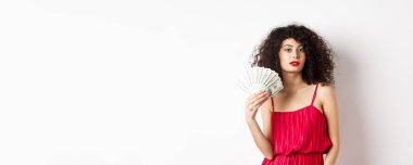 Zarif zengin kadın dolar banknotlarını hayranı olarak kullanıyor, para gösteriyor ve kendinden emin görünüyor, kırmızı elbiseyle beyaz arka planda duruyor..