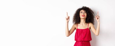 Kırmızı elbiseli hayalperest genç kadın, yukarı bakıyor ve logoyu işaret ediyor, mutlu gülümsüyor, beyaz arka planda duruyor. Boşluğu kopyala