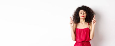 Kırmızı elbiseli, umut dolu bir kadın, dua ediyor, parmakları çapraz ve kapalı dua ediyor, tanrıya yalvarıyor, beyaz arka planda duruyor..