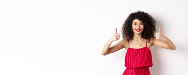 Kıvırcık saçlı, kırmızı elbiseli, parlak makyajlı, onay veren güzel bir kadın manken, evet de, gülümse ve başını salla, beyaz arka plan..