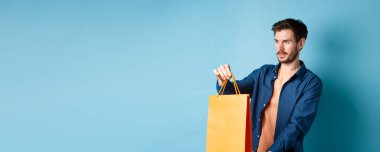 Yakışıklı genç adam, alışveriş satıcısı müşteriye turuncu alışveriş torbası uzatıyor, yan bakıyor, mavi arka planda duruyor..