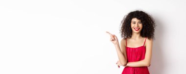 Kıvırcık saçlı güzel bir kadın, şenlikli bir elbise giyiyor, sol elini logoya gösteriyor ve gülümsüyor, beyaz arka planı var..