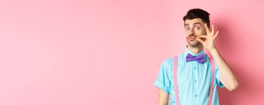 Komik genç adam Fransız bıyığına ve büzüşmüş dudaklarına dokunuyor, kameraya bakıyor, papyon takıyor ve pembe arka planda pantolon askısı takıyor..