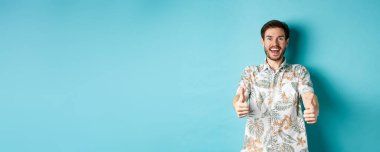 Havalı tebessüm eden, Hawaii tişörtlü adam aferin, şaşkın görünüyor, havalı bir tanıtıma bakıyor, mavi arka planda duruyor..