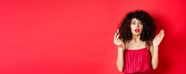 Güzellik ve makyaj konsepti. Kıvırcık saçlı heyecanlı bir kadın, kameraya bakarken nefesi kesiliyor, kırmızı ruj tutuyor, beyaz arka planda elbise giyiyor..