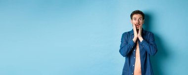 Şok olmuş yüzüyle soluk soluğa kalan, kameraya bakan, mavi arka planda duran adamın portresi..