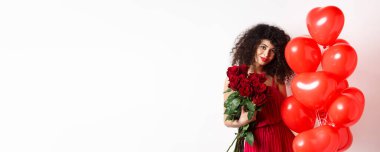 Gece elbisesi içinde güzel kıvırcık saçlı kız arkadaş, randevusu var, erkek arkadaşından kırmızı güller tutuyor ve romantik kalp balonlarının yanında poz veriyor, beyaz arka plan.