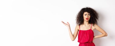 Üzgün, kıvırcık saçlı, kırmızı elbiseli, elini kaldıran, kafası karışmış, kaşlarını çatmış, haksız şeylerden bahseden, beyaz arka planda duran güzel bir kadın..